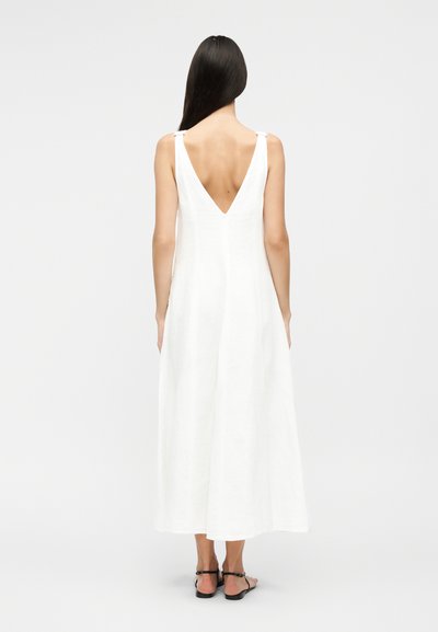 Hosszú sötét hajú nő, fehér ujjatlan ruhát visel mélyen kivágott hátul, fekete lapos szandálokkal, háttal áll fehér háttér előtt.