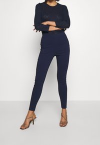 Tmavě modré přiléhavé kalhoty s vysokým pasem a hladkou texturou; doplněné páskovými sandály na vysokém podpatku s jemným hadím vzorem.