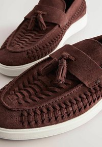 Chaussures slip-on en suède marron avec des détails tissés, semelle blanche plate et pompons décoratifs sur une sangle au-dessus.