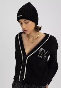 Czarny dzianinowy kardigan z białym wykończeniem, głębokim dekoltem w serek i czarnymi guzikami. Posiada wyszywane logo 'M'. Połączony z czarną, prążkowaną czapką.