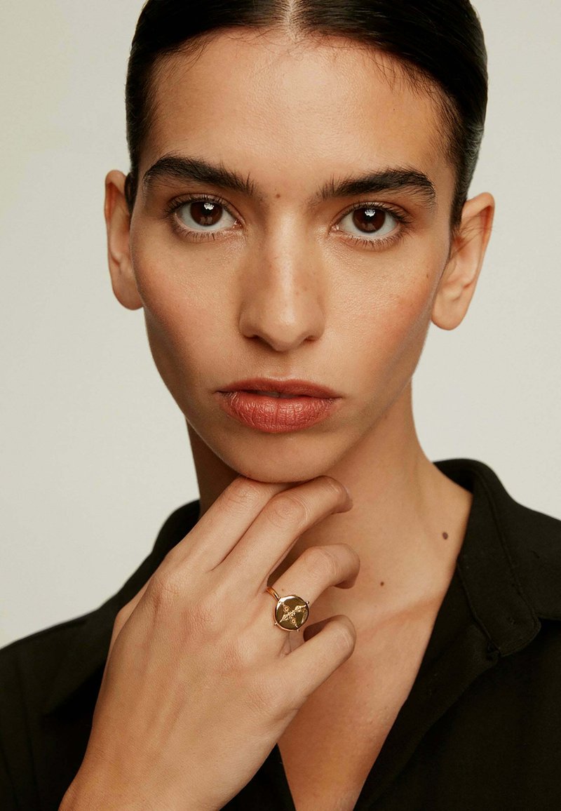 Anillo de oro con una gran piedra ovalada que presenta un patrón detallado. Mano del modelo cerca de la cara, vistiendo una camisa negra con cuello.
