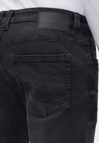 Κοντινό πλάνο της πίσω τσέπης μαύρου τζιν με ραμμένο σχέδιο και μαύρη δερμάτινη ετικέτα της επωνυμίας Tom Tailor στη ζώνη της μέσης.