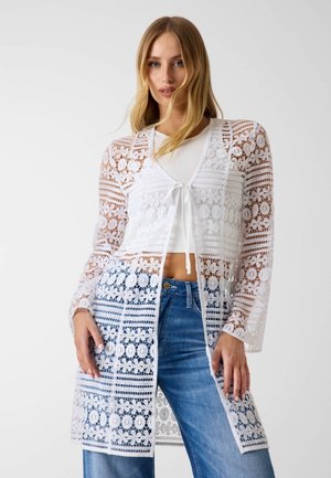 Femme portant un long cardigan en dentelle florale blanche par-dessus un crop top blanc et un jean bleu, debout devant un fond uni.