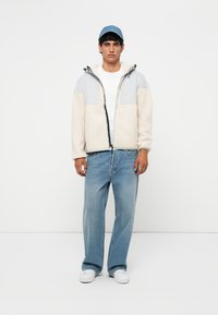 Light-colored fleece jacket with gray accents, zip closure, and two front pockets. Worn over a white t-shirt and paired with light blue jeans.