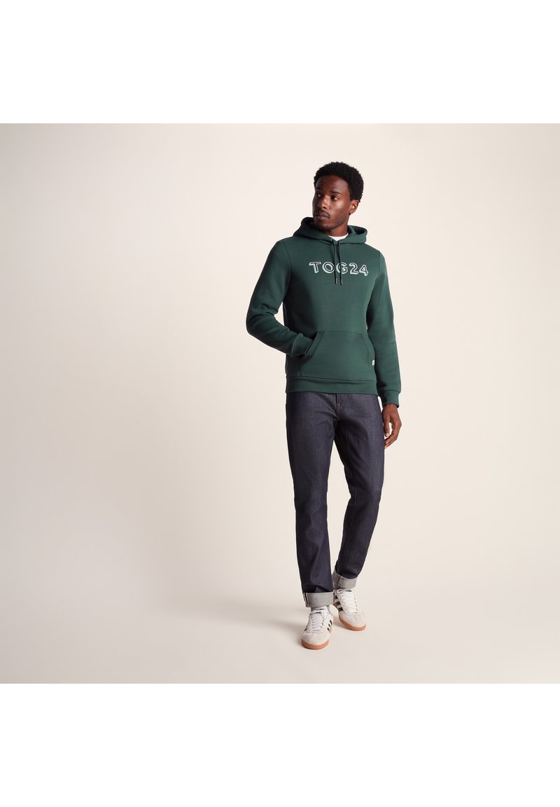 Green hooded sweatshirt with white lettering, front kangaroo pocket, paired with dark denim jeans and light shoes, on a neutral background.