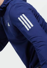 Mornarsko plava sportska hoodie s dugim rukavima, s bijelim Adidas trostrikama logom na lijevoj ruci i glatkom, teksturiranom tkaninom.