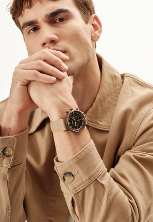 Ung mand i beige jakke, der hviler hagen på sammenfoldede hænder, iført et sort urskive-armbåndsur med beige stofrem.