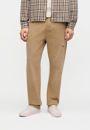 Mann trägt beige, ausgewaschene Jeans mit geradem Bein, weiße Turnschuhe und ein beiges kariertes Hemd zum Knöpfen.