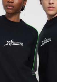 Δύο άτομα φορούν μαύρα φούτερ με το λευκό λογότυπο αστεριού "COSMO" στο στήθος, σε απλό λευκό φόντο.