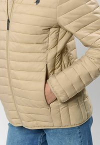 Beigas izolēta jaka ar horizontālām šuvēm, priekšējām kabatām un rāvējslēdzēja aizdari. Uz krūtīm ir mazs izšuvums ar logotipu.