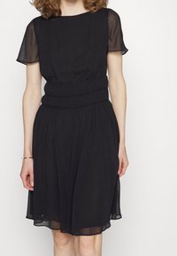 Černé šaty s krátkými průsvitnými rukávy, upnutým pasem a volnou sukní. Hladký materiál s jemným leskem a subtilními sklady.