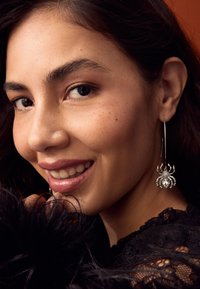 Silver spider earrings with intricate details and gemstones, worn by a woman with dark, wavy hair against a dark textured background.