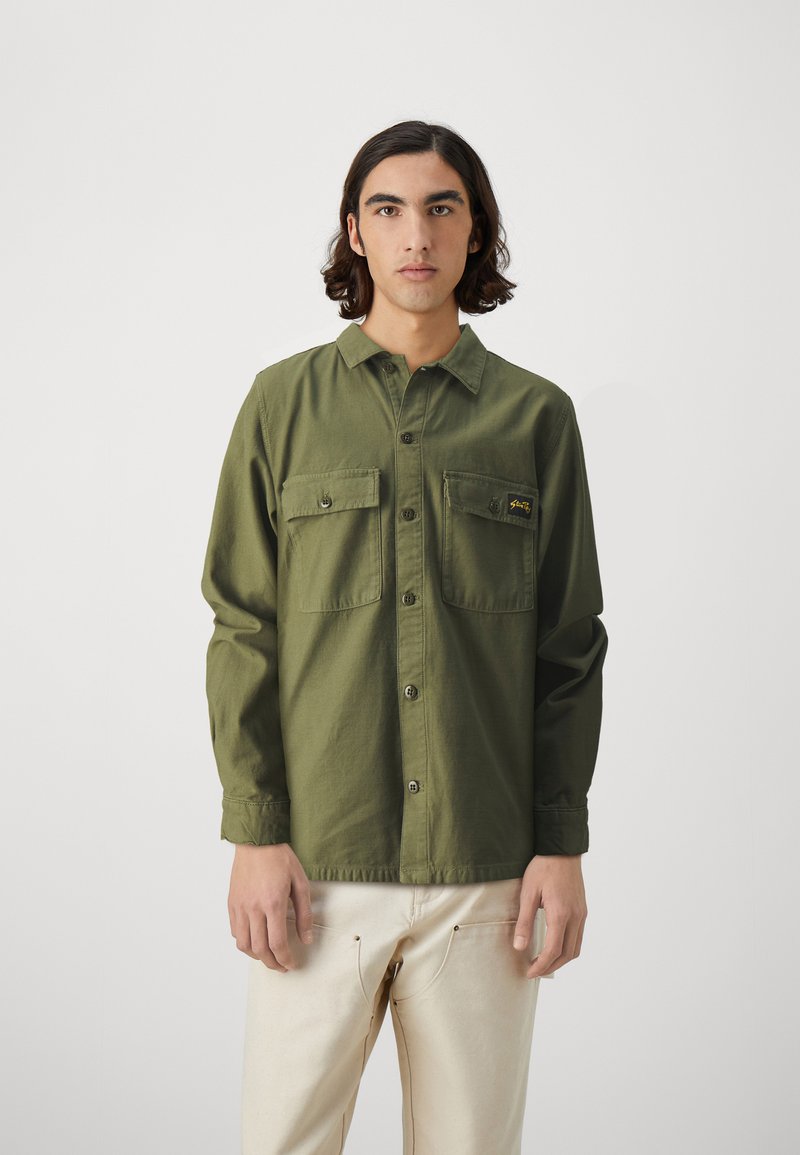 Green long-sleeve shirt with two chest pockets, button closure, and a small logo on the pocket. Smooth fabric, relaxed fit, and cuffed sleeves.