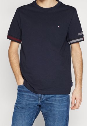 Mężczyzna ubrany w granatowy T-shirt z małym logo flagi na piersi, w paskowane ściągacze na rękawach i niebieskie dżinsy, stojący z jedną ręką w kieszeni.