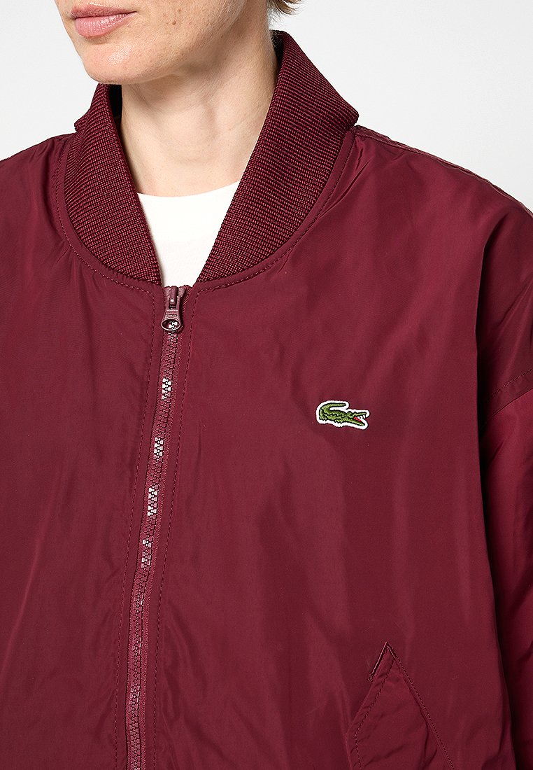 Veste bomber bourgogne avec un col côtelé texturé, fermeture éclair à l'avant et logo crocodile vert brodé sur la poitrine.