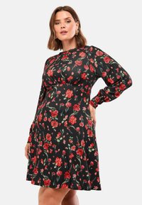 Juodas gėlių suknelė su raudonais ir žaliais gėlių raštais, ilgos rankovės, susiraukšlėjęs liemuo ir išplatėjusi sijonas. Glotnus audinio tekstūra, kasdieniškas dizainas.