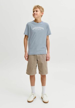 Dreng med kort blondt hår iført en lyseblå Jack & Jones t-shirt, beige shorts, hvide sokker og hvide sneakers, står mod en neutral baggrund.