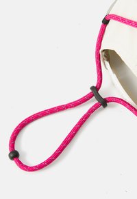 Corda intrecciata pink regolabile con un cursore in plastica nera, attaccata a un flap di tessuto chiaro con un bordo curvo.