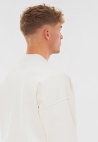 Bílá mikina se klasickým kulatým výstřihem, měkkou texturou, spadlými ramenními švy a minimalistickým designem. Viditelné švy na rukávech.