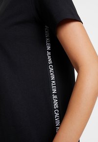 Czarna bawełniana koszulka z krótkim rękawem, ozdobiona białą taśmą z logo na boku z napisem "CALVIN KLEIN JEANS."