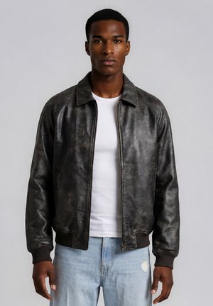 Hombre con una chaqueta de cuero negra sobre una camiseta blanca y vaqueros azul claro, de pie frente a un fondo gris liso.