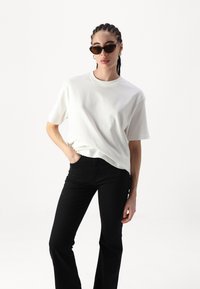 T-shirt de algodão branca com mangas curtas, corte descontraído e decote redondo, combinada com calças de ganga pretas com boca de sino, usadas com óculos de sol escuros.