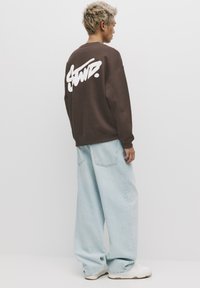 Rjava jopa z veliko belo grafiko na hrbtu, kombinirana s svetlo modrimi širokimi kavbojkami in belimi supergami. Gladek tekstilni material.