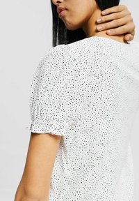 Blusa blanca con estampado de lunares negros, mangas cortas abullonadas con bordes con volantes y un corte relajado. Tejido suave con un leve brillo.