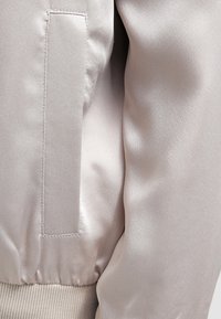 Veste bomber en satin beige clair avec une poche latérale, une taille côtelée et une texture lisse, mettant en avant un design moderne et épuré.