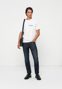 Hvid bomulds T-shirt med "Calvin Klein" logo, blå denim jeans og sorte lædersko. Modellen holder en lille sort crossover taske.