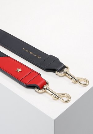 Deux anses de sac à main, une bleue marine avec un texte doré "Tommy Hilfiger", une rouge avec une étoile dorée, chacune avec des fermoirs métalliques dorés sur une surface blanche.