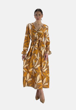 Femme portant une robe à manches longues, longueur cheville, avec un grand motif floral orange, beige et marron, taille nouée et talons beiges, debout face à l'avant.