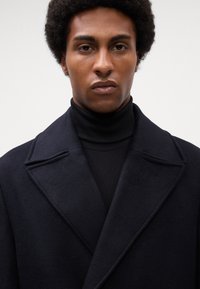 Man met afro-haarstijl, gekleed in een zwarte coltrui en een donkerblauw wollen jas, kijkt recht vooruit met een neutrale uitdrukking.