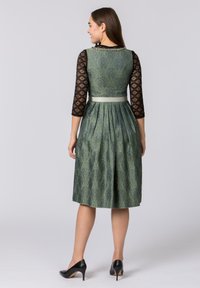 Grün gemustertes Kleid mit schwarzen Spitzen-Dreiviertelärmeln, plissiertem Rock und einem leichten Gürtelakzent an der Taille. Schwarze High-Heels.