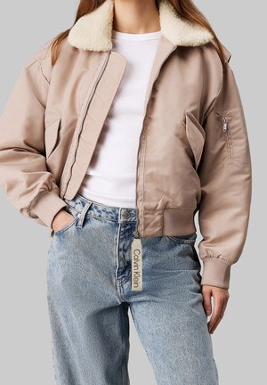 Frau trägt eine beige Bomberjacke mit weißem Fleecekkragen, weißes geripptes Shirt und hellblaue hoch taillierte Jeans mit Calvin Klein Reißverschluss-Tag.