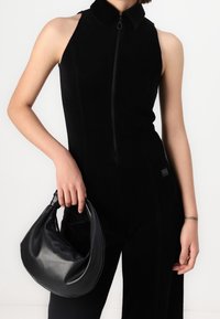 Mono negro con collar y cremallera frontal, hecho de tela texturizada. El modelo sostiene un bolso hobo de cuero negro con un asa anudada.