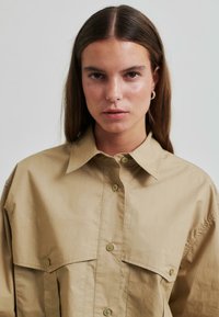 Cămașă supradimensionată kaki cu guler ascuțit, două buzunare frontale și închidere cu nasturi. Textură netedă, design casual cu o croială relaxată.