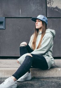Light green hoodie, black distressed jeans, and white sneakers. The cap is blue with an emblem. The subject sits on a concrete step.