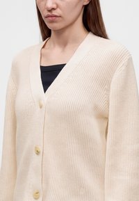 Jeune personne portant un cardigan en tricot couleur crème avec des boutons sur un haut noir, montrant le haut du torse et la zone du cou.