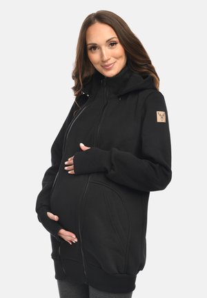 Pregnant woman gently holding belly, wearing black zip-up hoodie with thumb holes and a stitched deer logo on the sleeve.