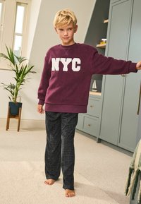 Tummanpunainen huppari, jossa on valkoiset "NYC"-kirjaimet, yhdistetty mustiin kuvioituihin pyjama-housuihin, esitelty kirkkaassa sisätilassa.