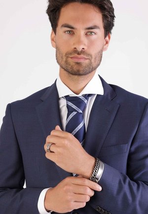 Veste de costume marine en tissu texturé, associée à une chemise blanche à col et une cravate rayée bleue et blanche. Bracelet en métal et bague visibles.