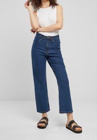 Jeans de mezclilla de cintura alta en azul oscuro, con un corte recto y un diseño de cinco bolsillos. Combinados con una blusa sin mangas blanca y sandalias negras.