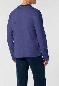 Uomo che indossa un pullover a maniche lunghe di colore blu scuro e pantaloni blu navy, in piedi leggermente girato con le mani rilassate ai fianchi.