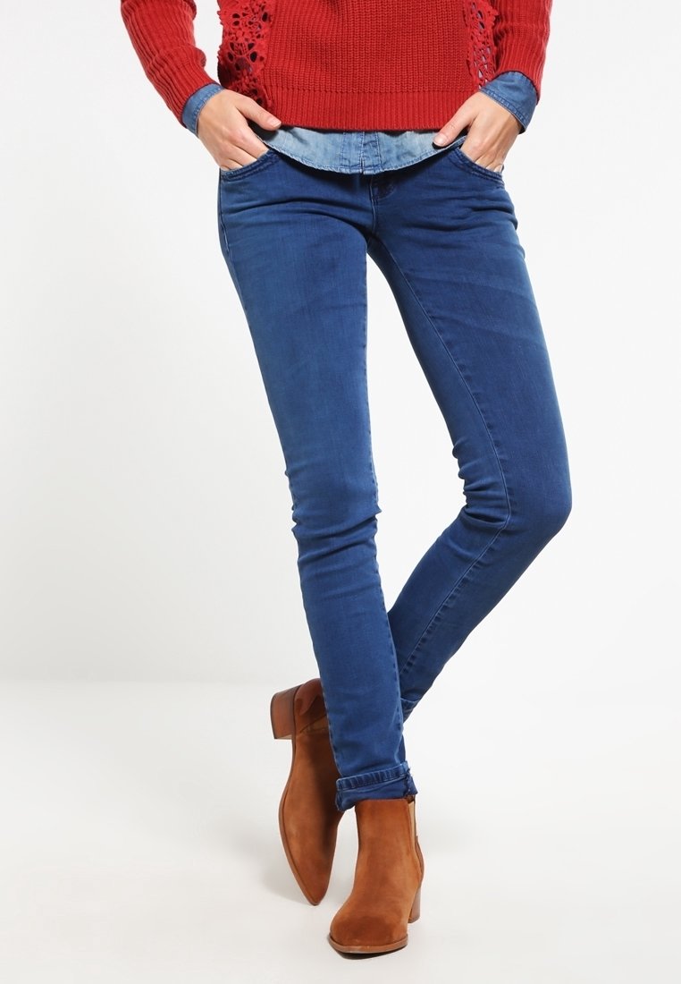 Jean en denim skinny bleu foncé, avec une texture lisse et une coupe ajustée. Associé à des bottines en daim marron.