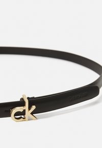 Ceinture en cuir noir avec une boucle logo "ck" en or. Texture lisse, design slim, avec un détail "Calvin Klein" en relief sur la sangle.