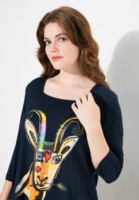 Laivansinisessä paidassa on neliön muotoinen pääntie ja kolmen neljänneksen hihattomat. Paidassa on värikäs vuohigrafiikka, jonka tehosteena ovat keltainen, punainen ja sininen.