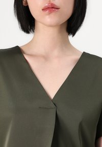 Blusa verde oliva con scollatura a V, caratterizzata da una superficie liscia e opaca e da dettagli di cucitura minimalisti sul davanti.