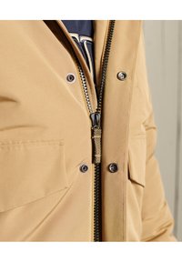 Chaqueta marrón con cierre frontal, botones a presión, dos bolsillos en el pecho y detalles en el cierre negro. Tejido suave con un acabado ligeramente brillante.