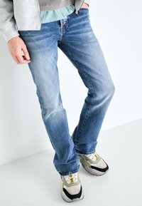 Jeans de mezclilla azul claro con corte recto, con un sutil desgaste en los muslos. Llevados con zapatillas deportivas grises y una camisa a rayas.